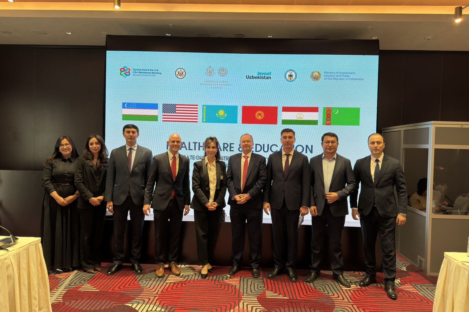 Nine delegates standing in front of the C5+1 Ministerial Meeting backdrop in Samarkand, which carries the flags of Uzbekistan, the United States, Kazakhstan, Kyrgyzstan, Tajikistan and Turkmenistan above the session title “Healthcare & Education”.
